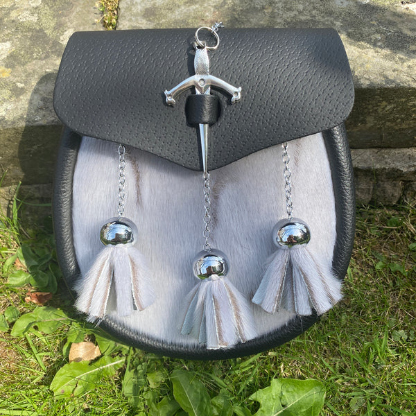 A black leather sporran with a light gray sealskin body, featuring a silver sword-shaped metal clasp and three hanging white and gray tassels.